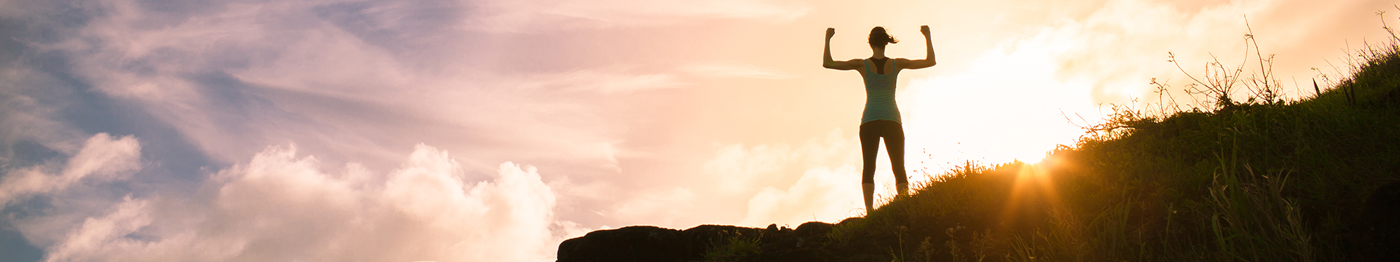 Image of woman standing triumphantly on a hill with her arms in the air looking into the sunset