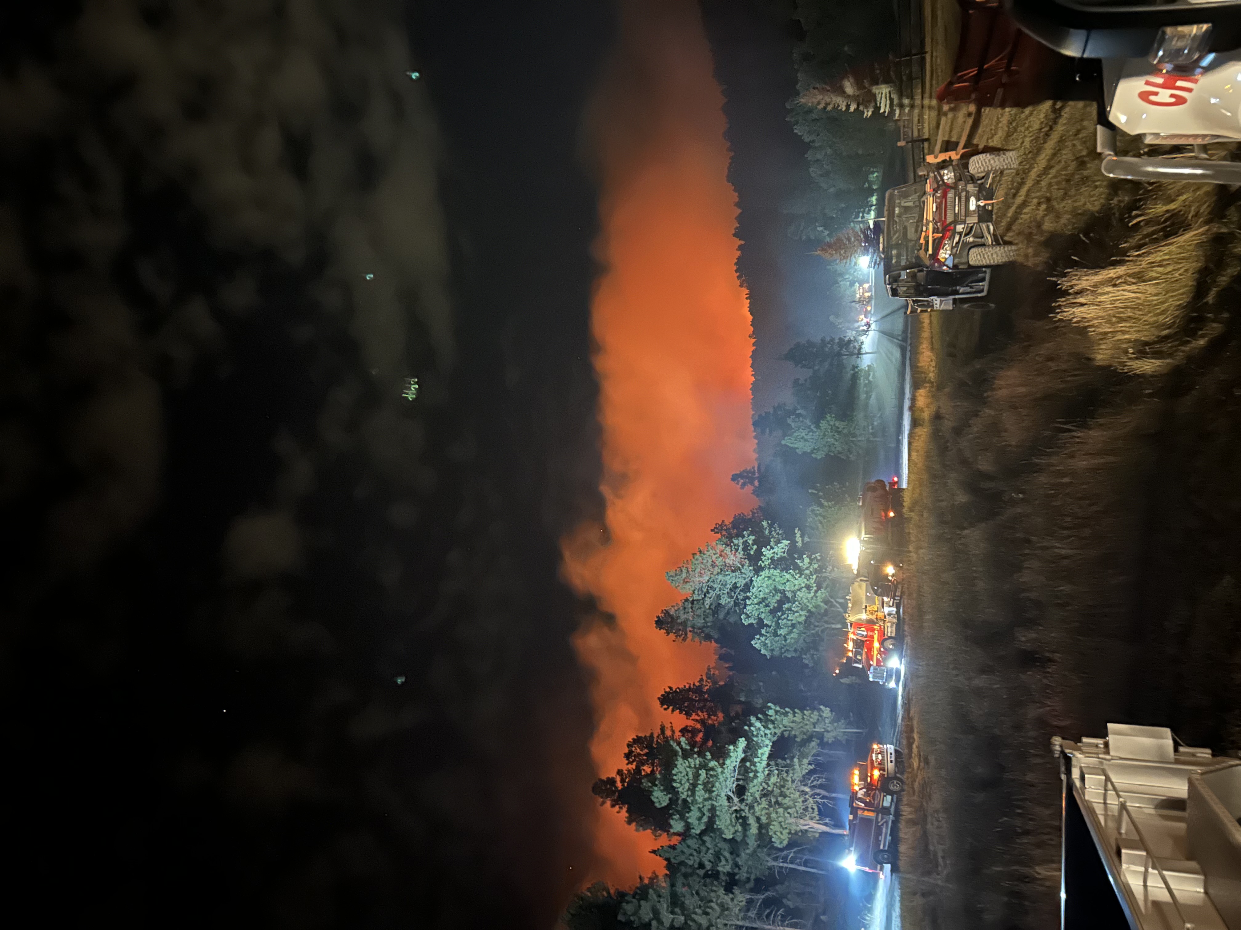 Image of nighttime forest fire with fire trucks and pine trees in the foreground and an orange glow coming from behind a hilltop