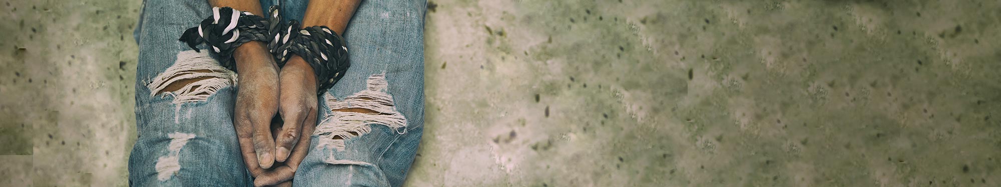 Image of person with ripped jeans sitting on the ground with their wrists on their knees tied in a rope